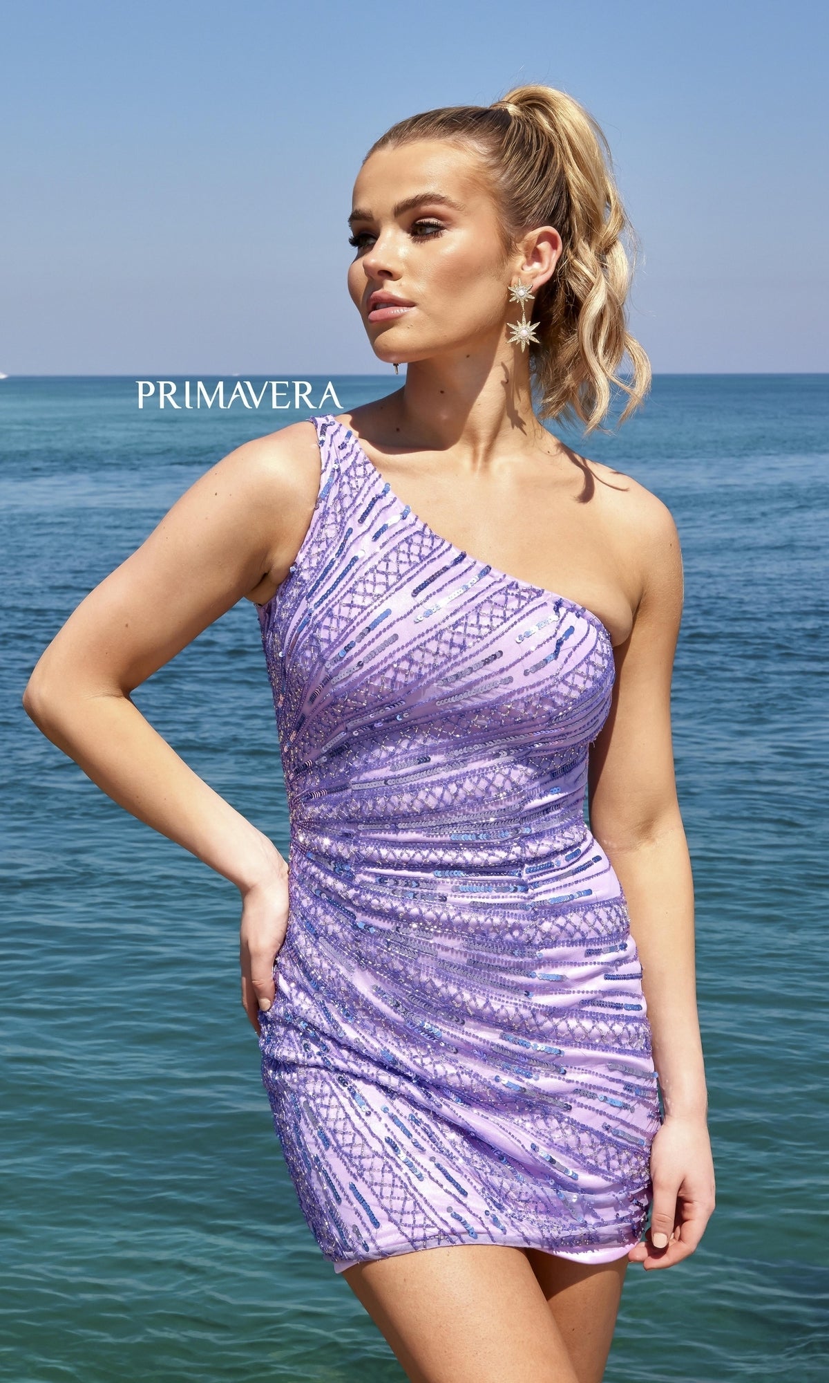 A woman stands by the ocean in the One-Shoulder Sequin-Striped Homecoming Dress 4046, a fitted light purple dress with sparkling geometric sequin patterns.